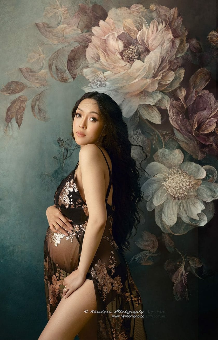 A radiant expectant mother dressed in a sophisticated black semi-sheer gown, standing before a charming floral Shades of Drops backdrop.