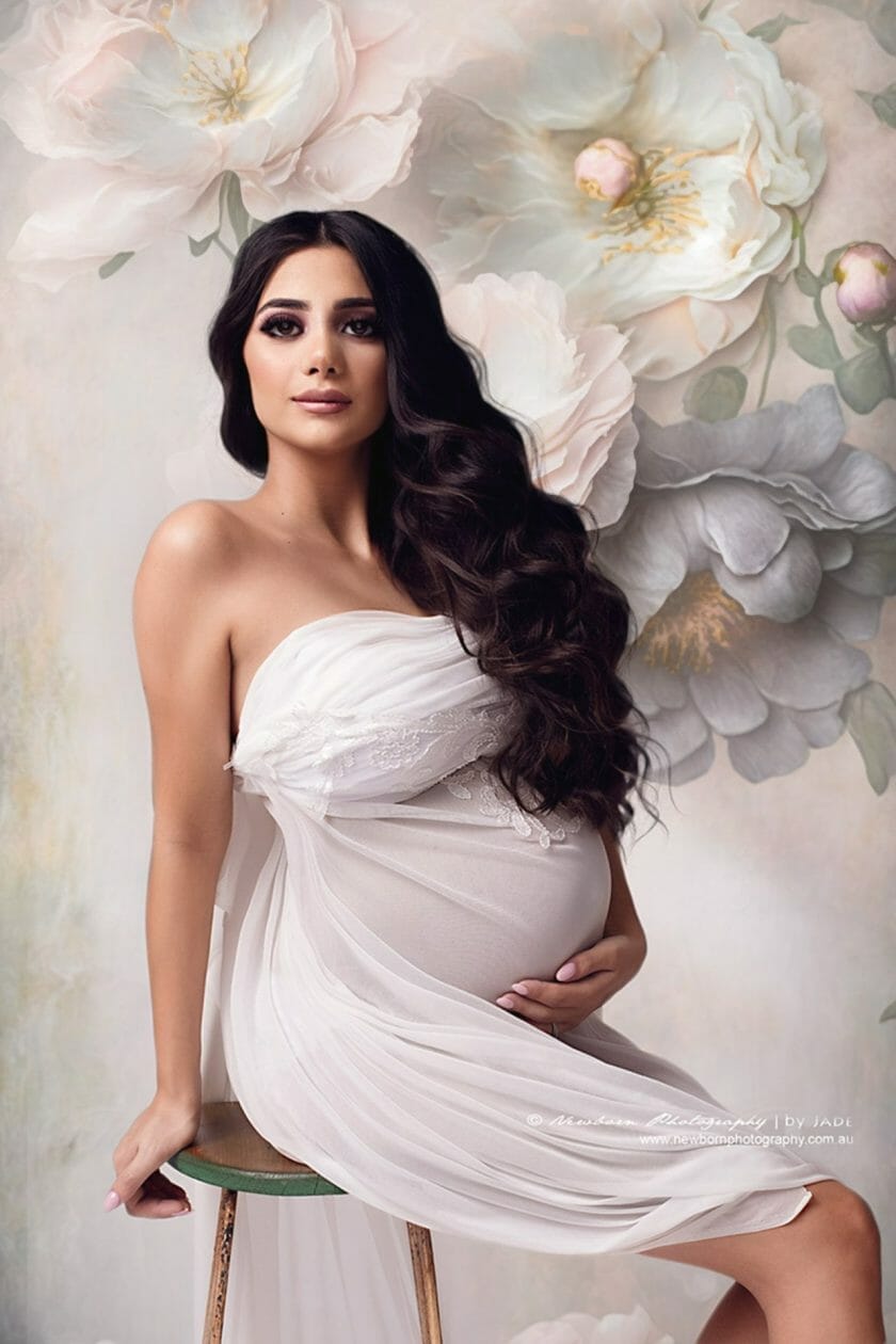 An elegant pregnant woman, adorned in a flowing white gown, stands in front of a backdrop decorated with artistic droplet designs intermingled with floral motifs."